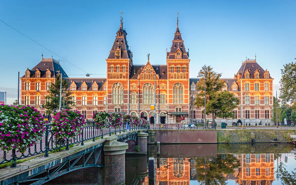 The grand façade of the Rijksmuseum in Amsterdam with tulip gardens and visitors walking through Museumplein under blue skies.